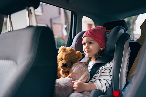 Cheerful girl with a red hat holding her favorite fluffy toys while sitting in car safety seat and looking in front of her.
