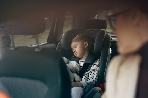 Cute little girl in a child safety seat napping with her toy in the car while her mother is driving. She is strapped in the seat and her head has fallen on the side.
