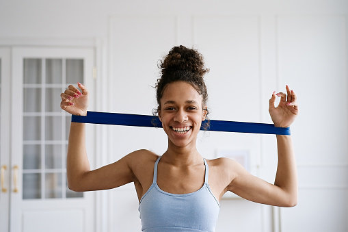 Home Sport. African american woman atching online tutorials on laptop and training on yoga mat in living room. Young girl exercising with fitness elastic bands and enjoying fitness and healthy lifesty