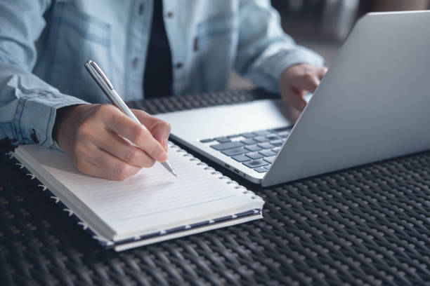 istock Online class, Student writing on notebook while study at home. Young adult woman doing online lesson by using laptop computer 1424230211