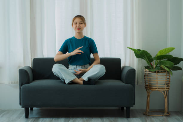 donna asiatica che medita, seduta sul divano, gambe incrociate, a piedi nudi piegati, medita, tranquilla facendo yoga nel soggiorno calmo yogi concetto di yoga e meditazione - yoga business women living room foto e immagini stock