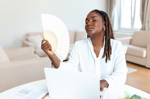 Tired African woman work on laptop at home office wave with hand fan suffer from heatstroke indoors. Unwell exhausted young female use waver breathe fresh air, lack ventilation conditioner.