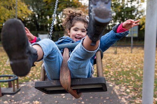 A young girl playing on a push swing at a public park in Hexham, North East England during Autumn. She has her arms outstretched while looking at the camera and smiling.