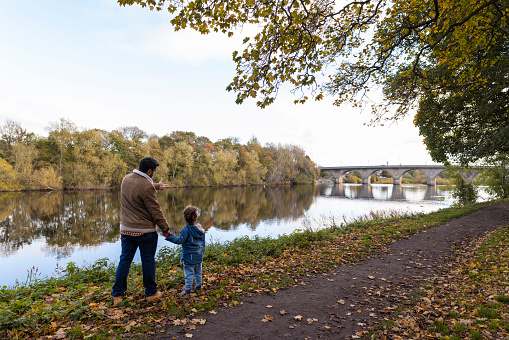 A man standing in a public park next to a river with his daughter in Hexham, North East England during Autumn. He is talking to her while pointing at the river.