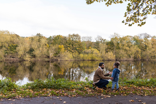 A man crouching down in a public park next to a river while talking to his daughter in Hexham, North East England during Autumn.