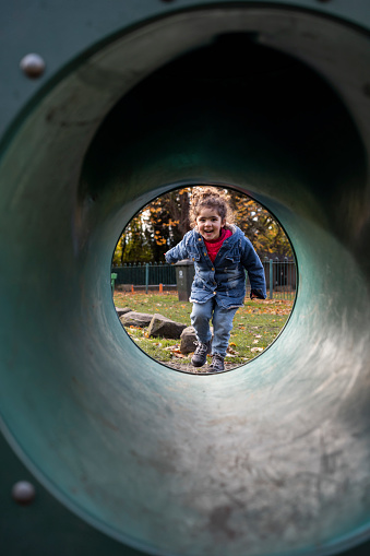 A young girl looking through a play tunnel at a public park in Hexham, North East England during Autumn. She is looking at the camera, about to run through the tunnel excitedly.