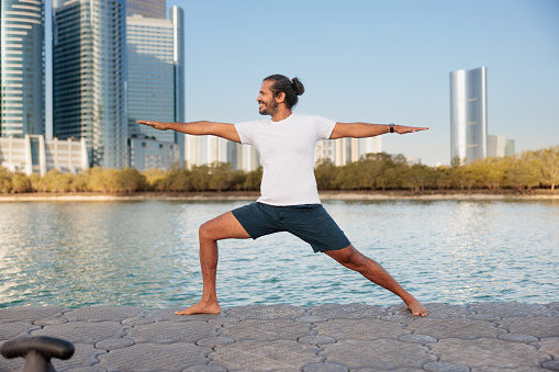 Man doing yoga on pier by the sea in Abu Dhabi, standing in lunge with arms outstretched in Warrior 2 Virabhadrasana II pose, cityscape in the background