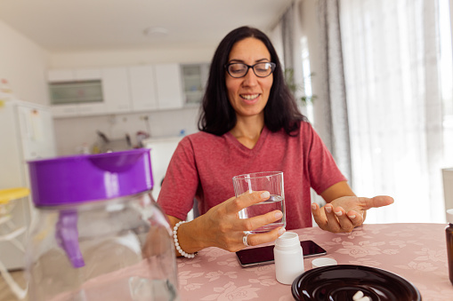A front view shot of a woman sitting at the dining table and taking vitamins with a glass of water at home.
