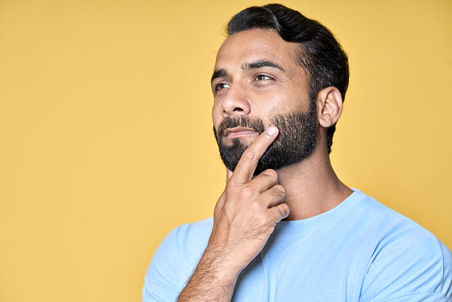 Thoughtful bearded indian man holding hand on chin looking interested aside at copy space isolated on yellow background thinking of new job opportunities, having doubt question or deciding concept.