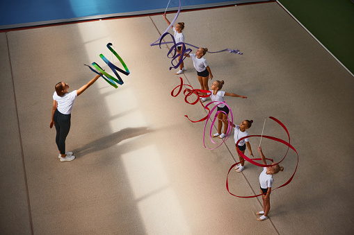 Ribbon exercises. Aerial view. Female sports coach training gymnastics athletes at sports gym, indoors. Concept of achievements, studying, goals. Coaching, training, child psychology