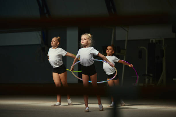 Flexible little girls, female rhythmic gymnasts training with gymnastic hoop at sport gym, indoors. Concept of action, motion, sport, motivation, competition. Beauty of movement. Flexible little girls, female rhythmic gymnasts training with gymnastic hoop at sport gym, indoors. Concept of action, motion, sport, motivation, competition. Group exercises coach bus stock pictures, royalty-free photos & images