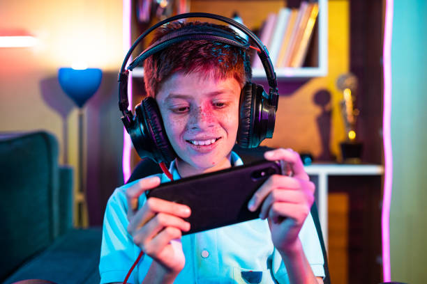 Happy teenager kid with headset playing video game on mobile phone at home - concept of relaxation, technology addiction and entertainment Happy teenager kid with headset playing video game on mobile phone at home - concept of relaxation, technology addiction and entertainment. digital native stock pictures, royalty-free photos & images