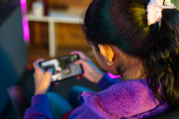 focus on girl, Shoulder shot of girl playing live video game on mobile phone at home - concept of relaxation, championship and tournament. focus on girl, Shoulder shot of girl playing live video game on mobile phone at home - concept of relaxation, championship and tournament bet live online stock pictures, royalty-free photos & images