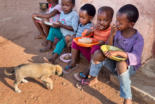 hungry African children eating porridge in front of the house in the village, puppy watching
