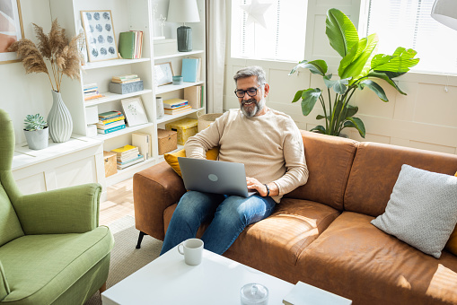 Mature man sitting on sofa at home and using laptop for business or pleasure.