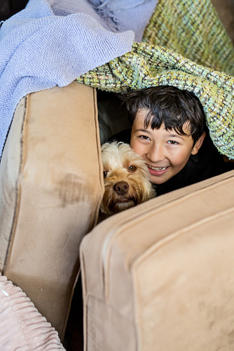 Boy and his dog sitting inside a pretend fort made of blankets and pillows looking up at the camera smiling and having fun together