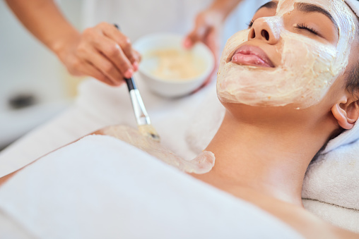 Facial treatment and spa beauty for a relaxed, calm and relaxing young woman doing skincare. Beautician applying a clay face mask or lotion on a happy Latino female enjoying the aesthetic procedure