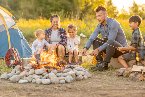 A small mixed race family sits closely around a fire as they roast marshmallows and enjoy time together.  They are each dressed comfortably and their tent can be seen set up in the background as they enjoy heir camping vacation.