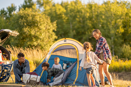 A small family is seen setting up their tent as they settle in to camp.  Their vehicle trunk is open as they unload and set up their campsite.  They are each dressed casually and focused on working together.