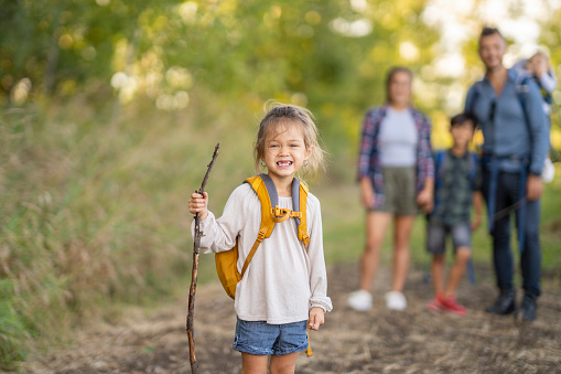 A sweet little girl pauses to pose for a photo during a family hike.  She is dressed casually, with a backpack on for her supplies and a walking stick in hand.  Her family can be seen standing in the background.