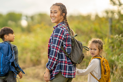 A young mother hikes with her two children on a warm summer day.  They are each dressed casually and have backpacks on with their supplies.  They are looking back to smile at the camera.