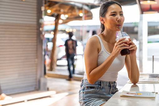 Attractive Asian girl drinking iced coffee at cafe in weekend market with smiling. Happy man and woman couple enjoy and fun outdoor lifestyle shopping together at street market on summer vacation.