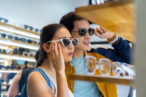 Young Asian couple enjoy and fun outdoor lifestyle shopping together at street market on summer holiday vacation. Happy man and woman couple choosing and buying fashion sunglasses together at fashion retail shop.