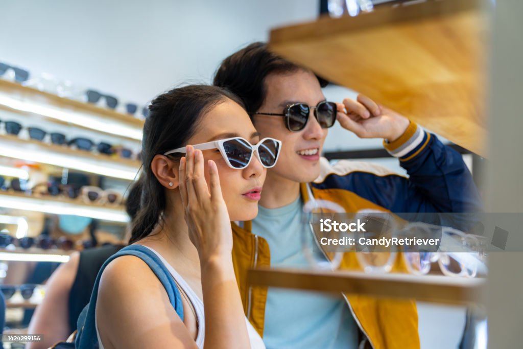 Young Asian couple shopping fashion sunglasses together at street market on summer vacation. Young Asian couple enjoy and fun outdoor lifestyle shopping together at street market on summer holiday vacation. Happy man and woman couple choosing and buying fashion sunglasses together at fashion retail shop. Shopping Stock Photo