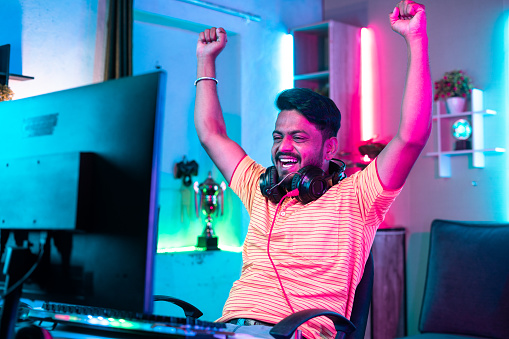 Cheerful gamer celebrating win by raising hands while playing online video game at home - concept of achievement, champion and successful.