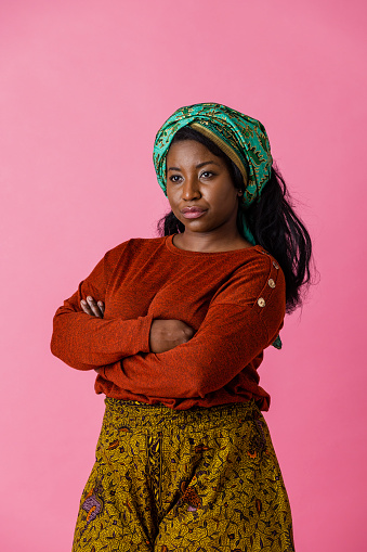 A studio portrait shot of a black woman wearing a head tie and casual clothing on a pink background. She is standing and looking away from the camera with her arms crossed and a serious expression on her face.