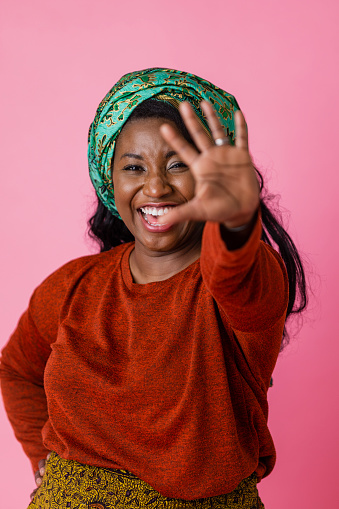 A studio portrait shot of a black woman wearing a head tie and casual clothing looking and smiling at the camera as she is holding one of her hands out towards the camera, on a pink background.