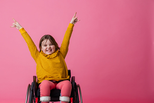 A studio portrait shot of a young girl who is using a wheelchair wearing casual clothing on a pink background. She is looking and smiling at the camera as she holds her hands in the air and makes rock hand gestures.