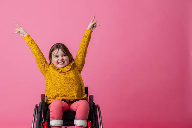 Rock Out! A studio portrait shot of a young girl who is using a wheelchair wearing casual clothing on a pink background. She is looking and smiling at the camera as she holds her hands in the air and makes rock hand gestures. special needs child pictures stock pictures, royalty-free photos & images