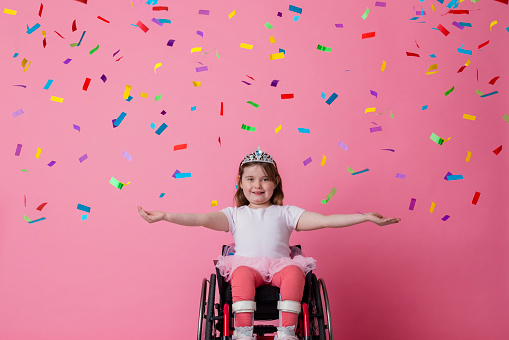 A studio portrait shot of a young girl who is using a wheelchair wearing ballerina clothing and a tiara on a pink background. She is holding her arms out by her side as she looks and smiles at the camera. Colourful confetti is falling around her.
