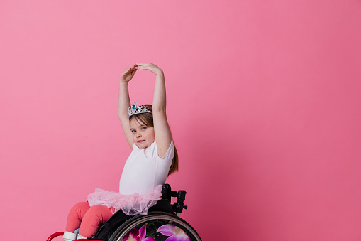 A studio portrait shot of a young girl who is using a wheelchair wearing ballerina clothing and a tiara on a pink background. She is doing ballet dancing as she looks at the camera.