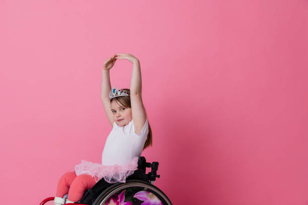 I Love Ballet A studio portrait shot of a young girl who is using a wheelchair wearing ballerina clothing and a tiara on a pink background. She is doing ballet dancing as she looks at the camera. special needs child pictures stock pictures, royalty-free photos & images