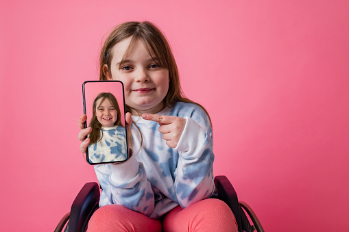 A studio portrait shot of a young girl who is using a wheelchair wearing casual clothing, on a pink background. She is holding out a smartphone to the camera with a photo of her on it.