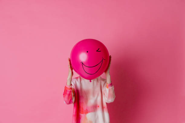 On a pink background is a three-quarter-length, studio portrait shot of a young woman wearing casual clothing. She is holding a pink balloon with a smile on the balloon in front of her face.