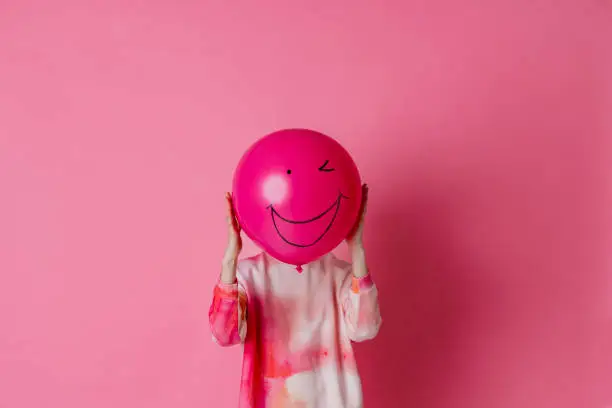 On a pink background is a three-quarter-length, studio portrait shot of a young woman wearing casual clothing. She is holding a pink balloon with a smile on the balloon in front of her face.