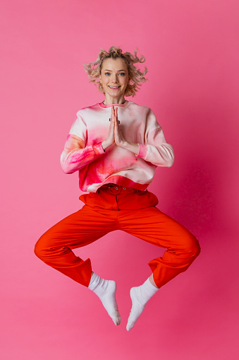 On a pink background, a full lenght studio portrait shot of a young woman wearing casual clothing looking into the camera jumping in the air with her legs out and her hands together.