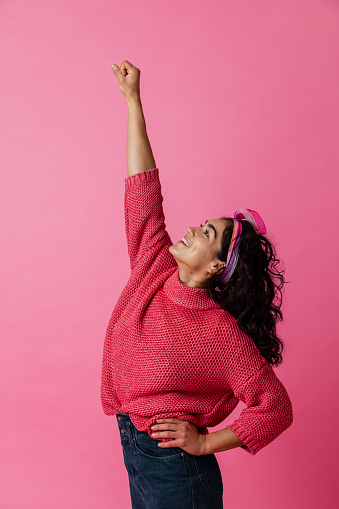 On a pink background, a three quarter length studio portrait shot of a young woman wearing casual clothing looking away from the camera as she poses like a superhero.