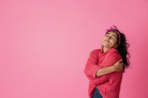 On a pink background, a three-quarter-length, studio portrait shot of a young woman wearing casual clothing embracing herself with her eyes closed, smiling.