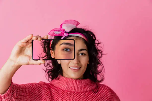 A studio portrait shot of a young woman wearing casual clothing, on a pink background. She is holding out a smartphone in front of her eye to the camera with a photo of her eye on it.