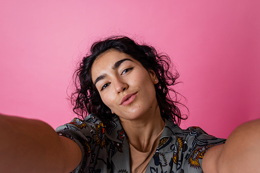 A studio portrait shot of a young woman wearing casual clothing, on a pink background. She is looking into the camera pouting with confidence as if she is taking a selfie.