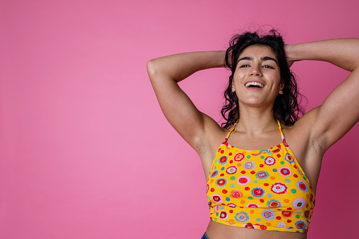 A studio portrait shot of a young woman wearing casual clothing, on a pink background. She has her hands in her hair with a positive emotion on her face.