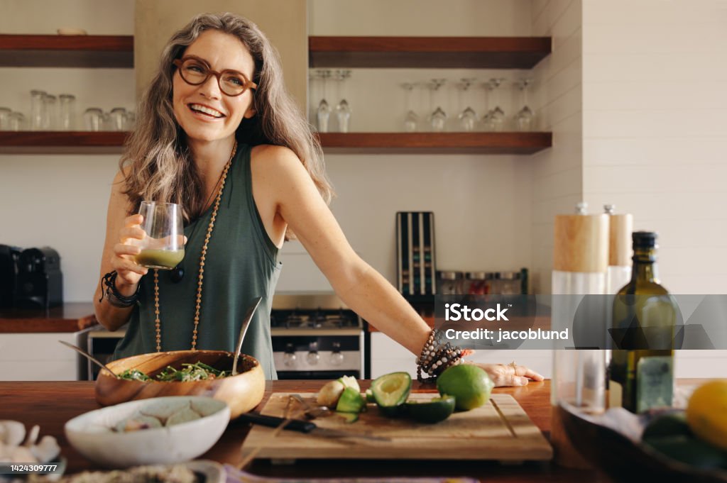 Healthy senior woman smiling while holding some green juice Healthy senior woman smiling while holding some green juice in her kitchen. Mature woman serving herself wholesome vegan food at home. Happy woman taking care of her aging body with a plant-based diet. Healthy Eating Stock Photo
