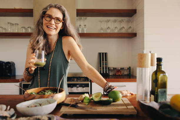 Healthy senior woman smiling while holding some green juice Healthy senior woman smiling while holding some green juice in her kitchen. Mature woman serving herself wholesome vegan food at home. Happy woman taking care of her aging body with a plant-based diet. cooking stock pictures, royalty-free photos & images