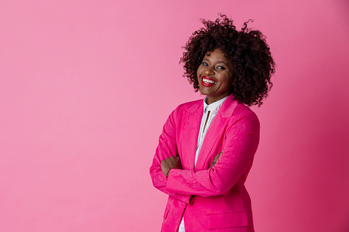 On a pink background, a studio portrait shot of a mature woman wearing smart matching workwear. She is looking into the camera smiling with confidence