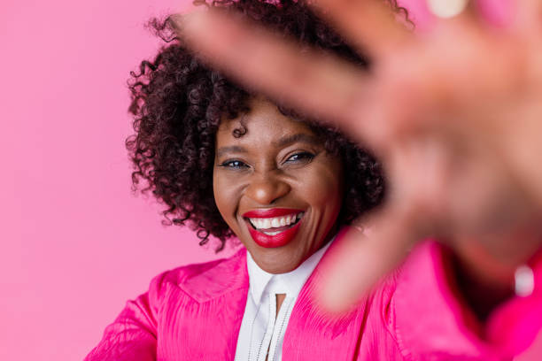 On a pink background, a studio portrait waist-up shot of a mature woman wearing smart workwear. She is looking into the camera with her hand up towards the camera smiling.