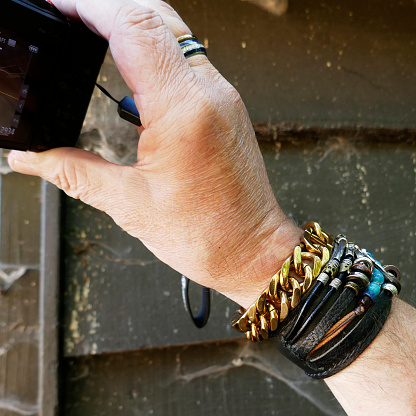 Senior man making a photo with a small camera. He is wearing a mixture of different bracelets. A large gold colored Cuban link bracelet, followed by a group of leather ones